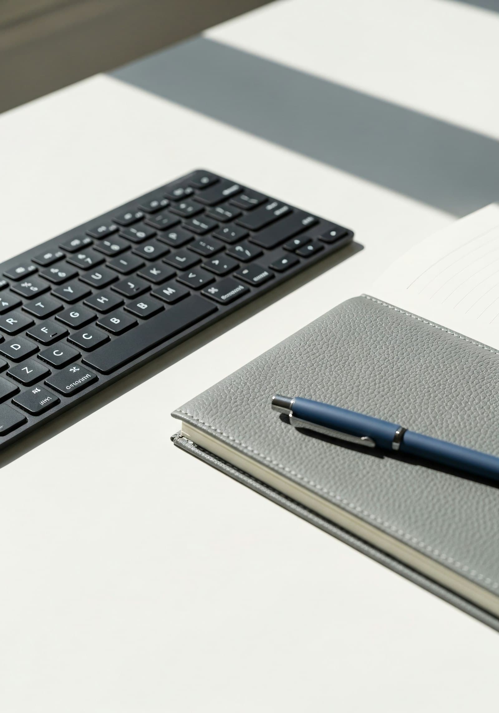 A notebook, pen and keyboard on a white desk in natural light